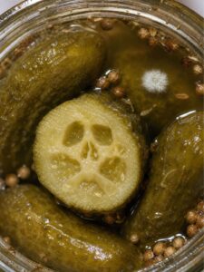 Fermented pickles in a jar, with white mold on the brine and a skull-like pattern inside a cut pickle. Photograph enhanced with AI-generated imagery.