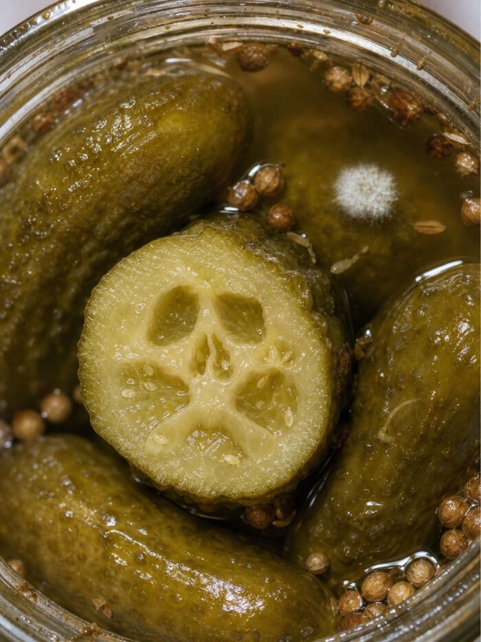 Fermented pickles in a jar, with white mold on the brine and a skull-like pattern inside a cut pickle. Photograph enhanced with AI-generated imagery.